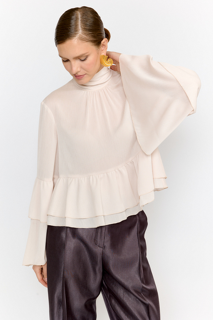 Блуза свободного кроя с легким расширением к низу с воланами и воротником-стойкой, переходящим в ленты, которые можно завязать в бант либо просто перекинуть через плечи. Выполнена из легкой полупрозрачной ткани. Крэш-шифон -легкий, дышащий материал, создающий эффект невесомости. Полиэстеровые волокна в составе обеспечивают   неприхотливость в уходе и устойчивость к механическим повреждениям. Нежный цвет экрю позволяет легко вписать блузу в гардероб, для создания как праздничных образов, так и повседневных.