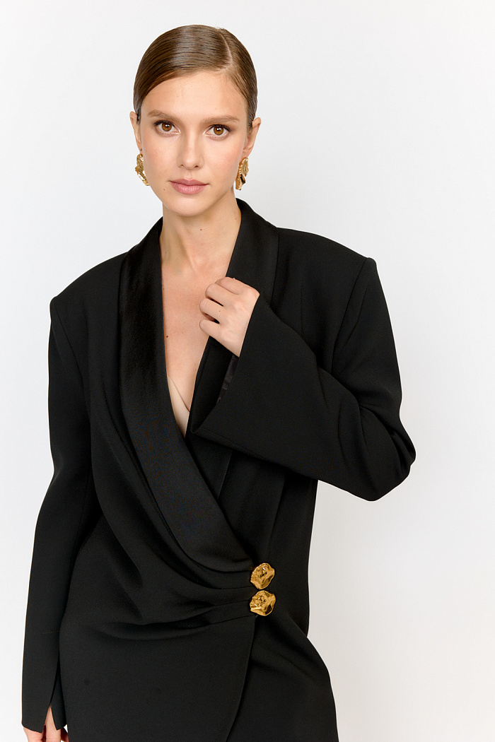 Жакет-смокинг прямого свободного кроя с расширенной линией плеча со складками по борту. Модель из костюмной ткани, основная часть которой с креповой, а воротник с атласной фактурой. По верхнему борту заложены три складки, застегивается на две декоративные пуговицы. Жакет выпилен из современной ацетатной ткани. Ацетатные волокна в составе придают блеск и  способность красиво драпироваться, обеспечивают неприхотливость в уходе, легкость, способность хорошо держать форму. Преимущества ткани:  имеет приятный блеск и мягкость, напоминающую натуральный шелк, но при этом выглядит более роскошно. Благодаря гибкости  и объемности, ткань красиво ложится по фигуре, создавая женственные силуэты.